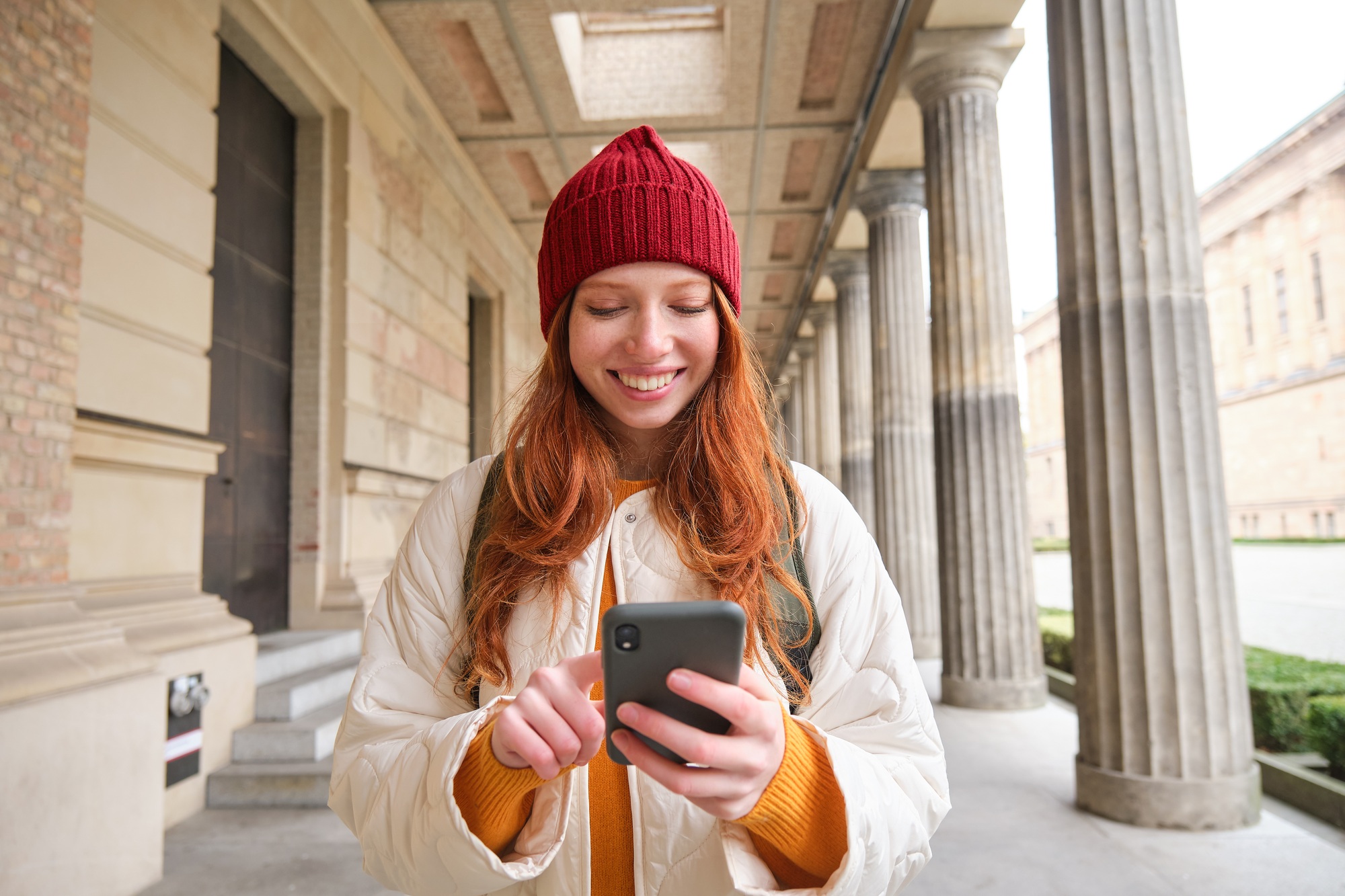 Smiling woman with red hair, tourist using mobile phone application while walking around town
